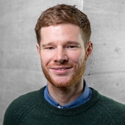 Man with red hair and beard, wearing a green sweater over a blue shirt, stands against a gray concrete wall, smiling.